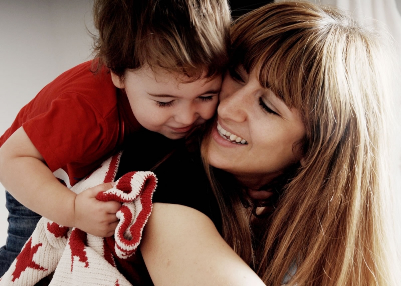 A mother and her toddler pressed cheek to cheek, both laughing, the little boy in a red top