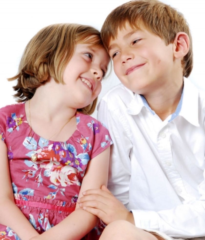 A young girl in a floral dress leaning against her older brother in a white shirt, both smiling