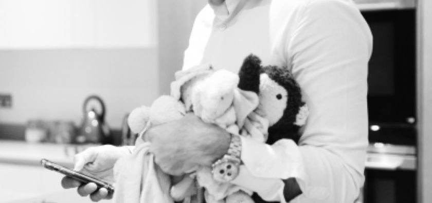 Black and white photograph of a father in his kitchen, cradling his baby wrapped in a blanket