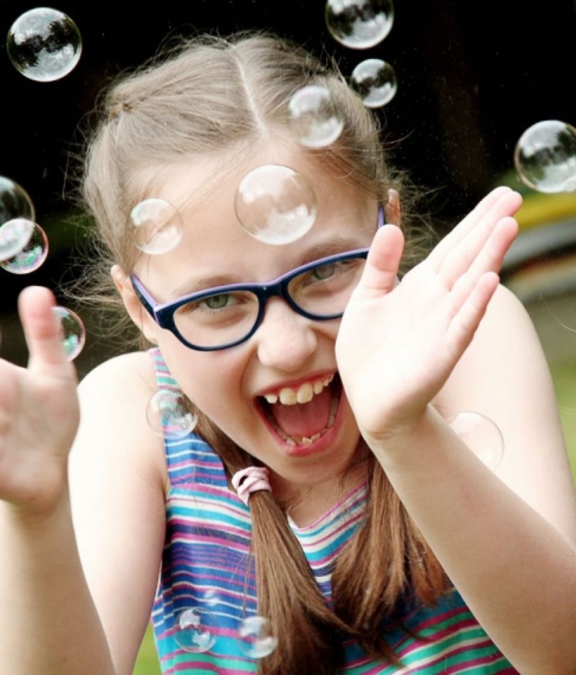 A girl in glasses laughing and reaching out as soap bubbles drift around her in a garden